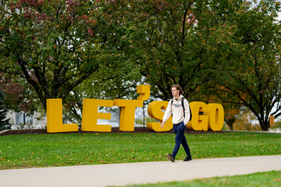 A students walks by giant gold letters that spell "Let's Go" on the Evansdale Campus.