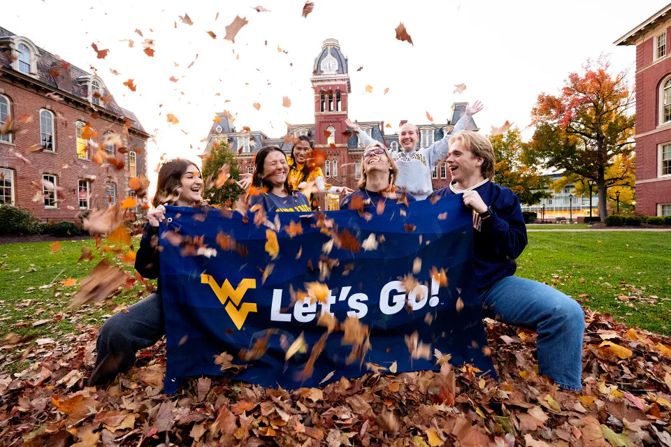 Four students hold a blue flag that says "Let's Go" while two students behind them throw fall leaves into the air.